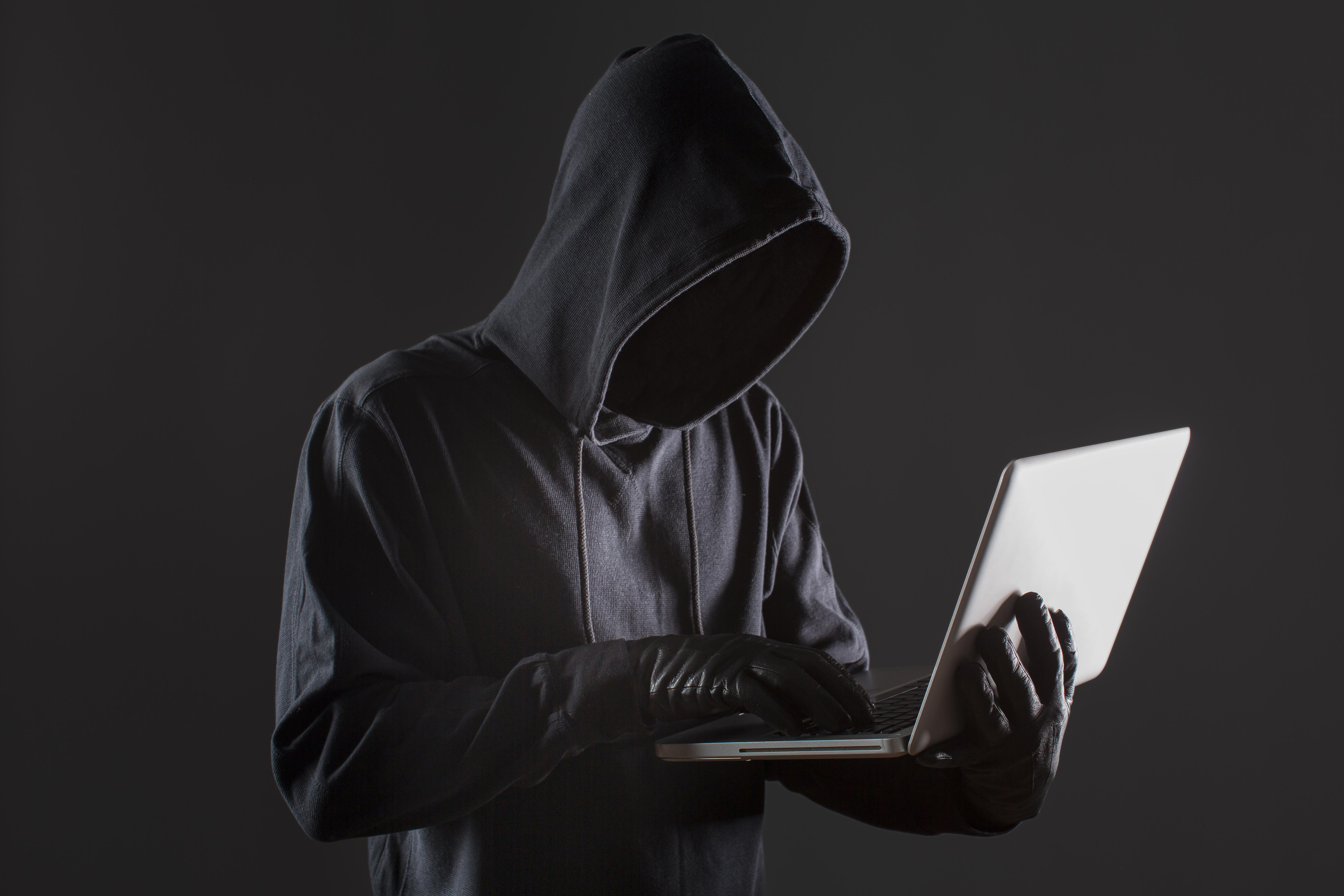 A side view of a suspicious man with a covered face and gloves using a laptop.