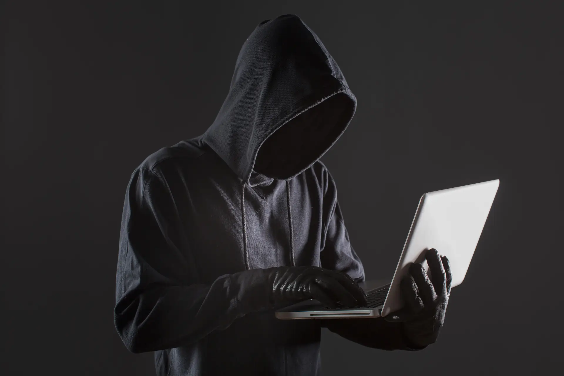 A side view of a suspicious man with a covered face and gloves using a laptop.